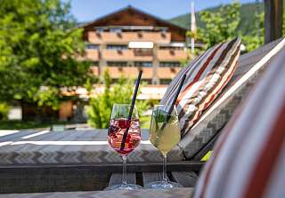 Zwei Sommerdrinks auf der Sonnenterrasse des Sunstar Hotels Klosters mit Blick auf das Hotelgebäude und die umliegende Berglandschaft.
