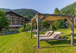 Hanging loungers under wooden pergolas in the garden of the Sunstar Hotel Klosters, with the hotel and forested mountains in the background in summer