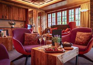 Cosy lounge at Sunstar Hotel Klosters with red armchairs, a wooden table with a platter of cured meats, bread basket and two glasses of sparkling wine, wood panelling and large windows behind.