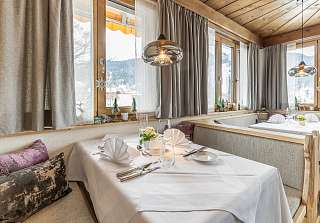 Laid table with upholstered bench and cushions by the window of the Bosca Stube at the Sunstar Hotel Klosters, with wooden ceiling, glass pendant lights and a view of the snowy landscape