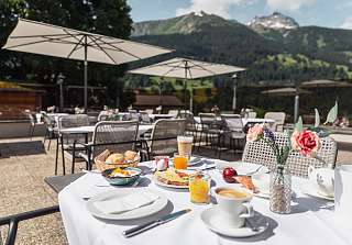 Table du petit-déjeuner dressée avec café, jus d'orange et bouquet de fleurs sur la terrasse ensoleillée du Sunstar Hotel Klosters avec panorama alpin