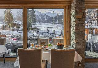 Table de petit-déjeuner dressée à la Bosca Stube du Sunstar Hotel Klosters, avec vue à travers les baies vitrées sur la terrasse enneigée et les montagnes